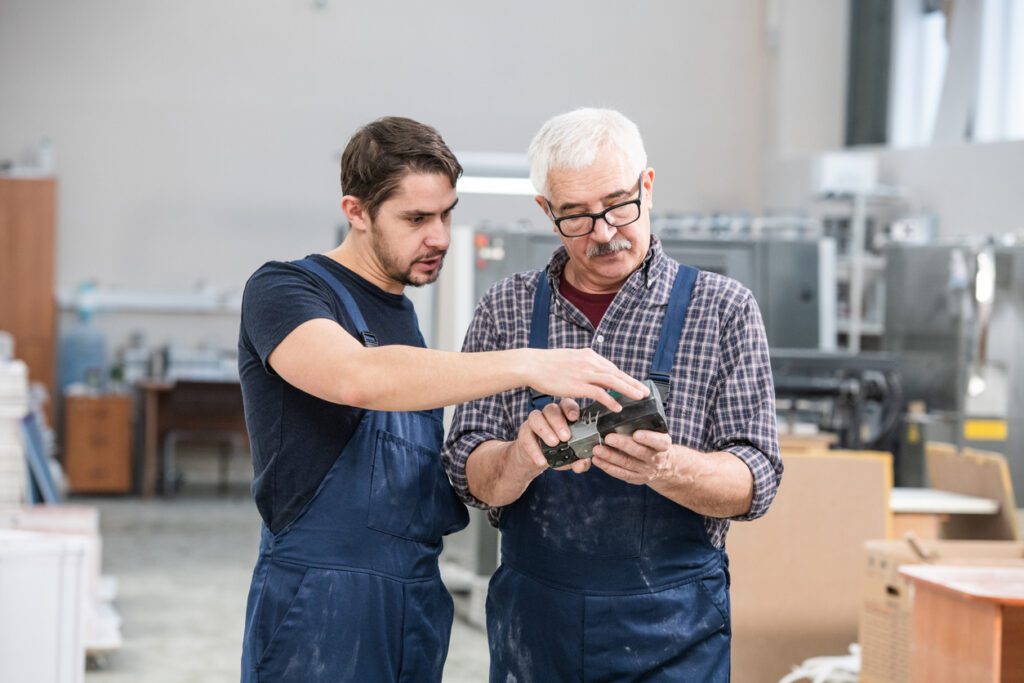 Young specialist in overall showing senior coworker how to use controller at factory