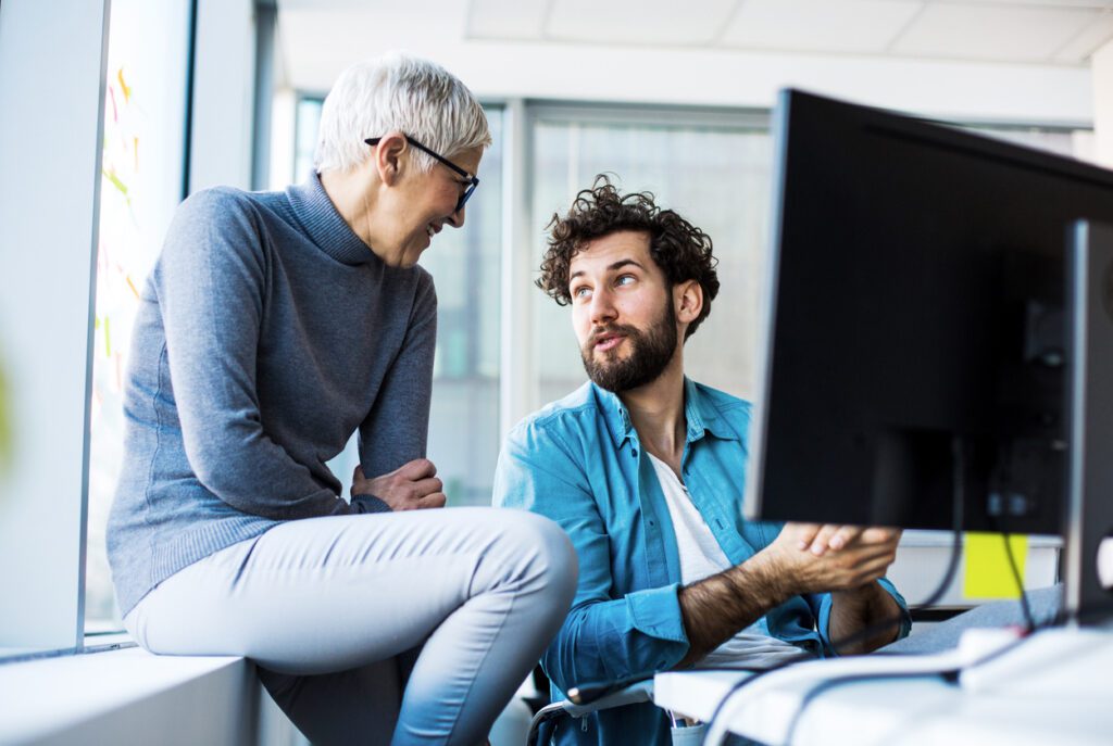 Two colleagues talking, thinking, smiling in their office. Younger business man is talking to senior colleague, feeling good about new job opportunities.