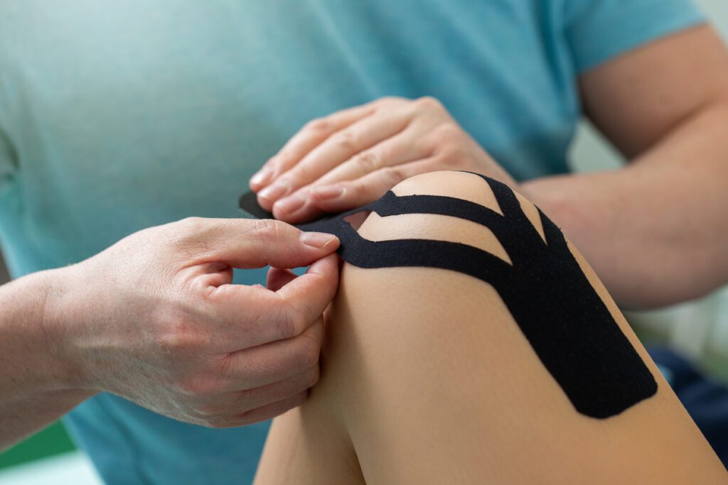Physiotherapist applying kinesiology tape to patient knee. Therapist treating female athlete with a functional bandage. Post traumatic rehabilitation and sport physical therapy.
