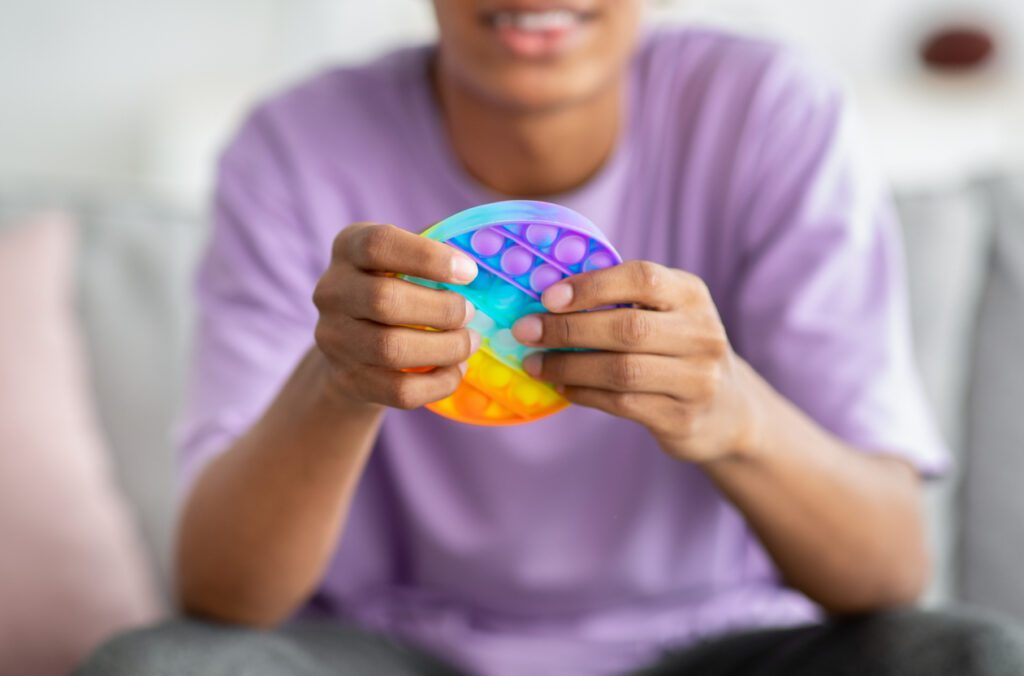 Closeup view of African American teenager playing with antistress POP IT toy at home. Unrecognizable black youth using sensory plaything to relieve stress or relax indoors