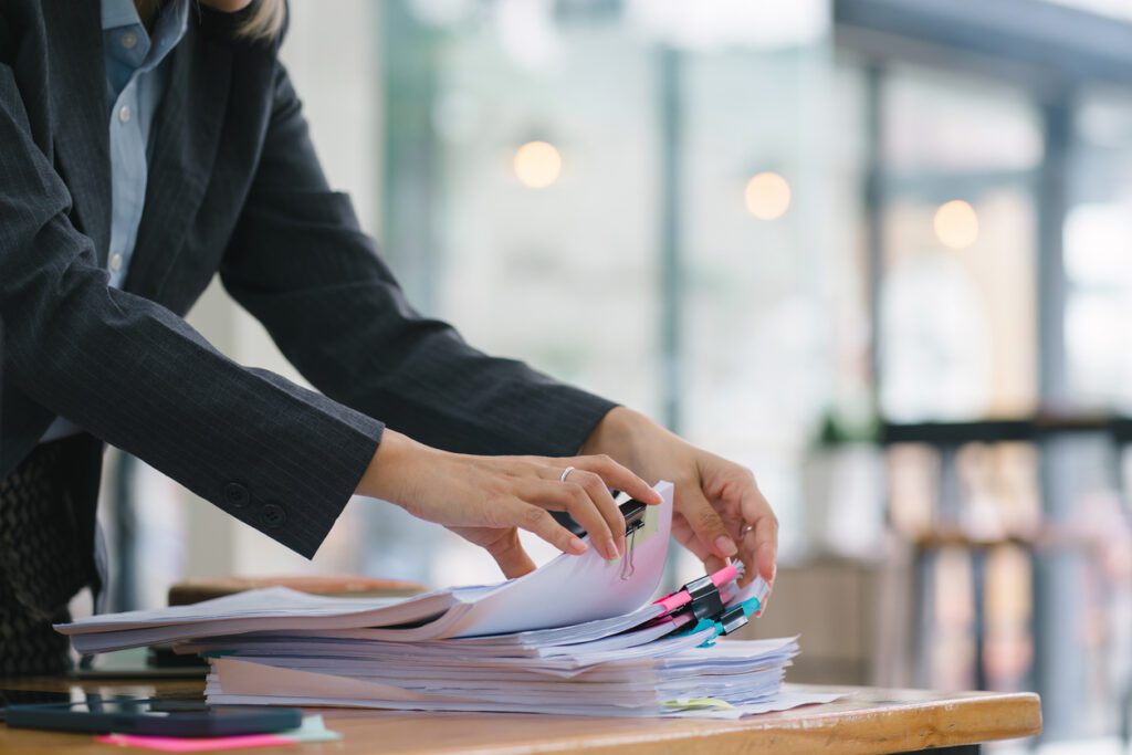 The businesswoman's hands are busy working amidst stacks of paper files, searching and checking for unfinished documents among the folders and papers on her cluttered office desk