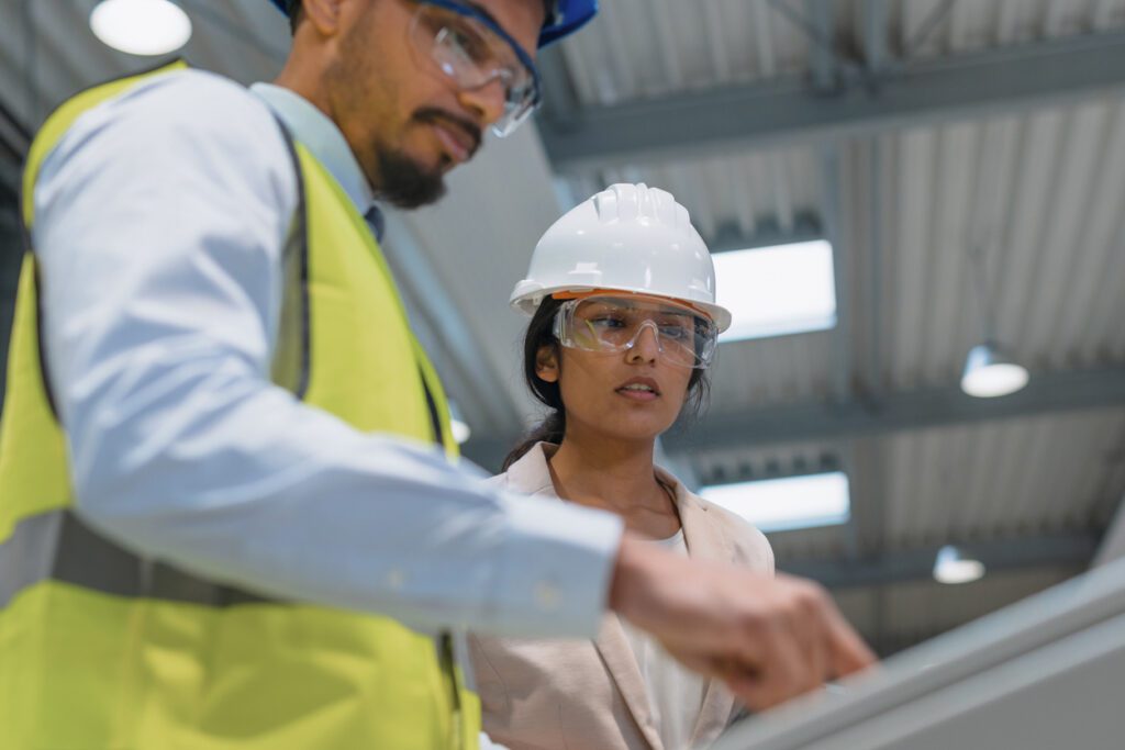Female Indian factory supervisor is on-the-job training an African American male employee, in a blue helmet and yellow vest, to operate machinery.